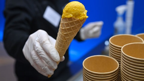 Eine Person hält in weißen Handschuhen Eiscreme in einer Waffel hoch. Daneben stehen aufgestapelte Eisbecher aus Pappe.
