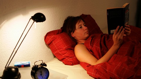 Eine junge braunhaarige Frau liegt abends im Bett und liest ein Buch.