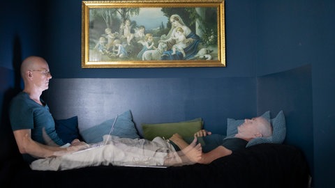 Ein Mann mit Glatze sitzt mit ausgestreckten Beinen auf der linken Seite mit Brille am Laptop auf der rechten Seite liegt dieser Mann mit geschlossenen Augen auf einem Kissen.