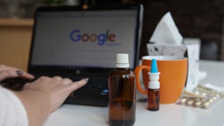 Eine Frau sucht etwas auf Google, auf dem Tisch stehen Medikamente | Wie hilfreich ist eine Diagnose von Dr. Google? Interview mit Ärzting Dr. Sibylle Siber
