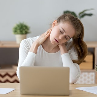Frau sitzt am Schreibtisch und fasst sich an den Nacken