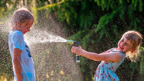 Abkühlung im Sommer: ein Mädchen bespritzt einen Jungen mit Wasser aus dem Gartenschlauch, beide lachen und freuen sich. Ab wann spricht man von einer Hitzewelle? Was ist der Hitzerekord in BW? Wie schütze ich mich am besten vor der Hitze? Der SWR1 Hitze-Ratgeber - mit einer interaktiven Wetterkarte für euren Ort, den schönsten Badespots (Flüsse, Seen, Hundestrände) in Baden-Württemberg und vielen weiteren Tipps, wie ihr euch bei Hitze selbst helfen könnt.