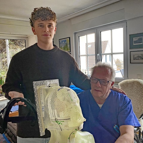 Ein junger Mann und sein Vater betrachten eine Gesichtsmaske zur Strahlentherapie aus gewebtem Kunststoff. SWR1 Neuanfang: Nachwuchsfußballer Max und sein Kampf gegen den Krebs. Bei dem jungen Mann aus Heidelberg wird ein Sarkon diagnostiziert, ein höchst aggressiver Krebs. Der Sport und seine Familie geben ihm die Kraft, die Chemotherapie und den Kampf gegen den Krebs erfolgreich zu bestehen.