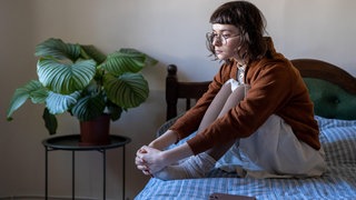 Eine Frau sitzt traurig mit den Knien angewinkelt alleine auf dem Bett | Einsamkeit und mentale Gesundheit: Warum Stress uns einsam macht