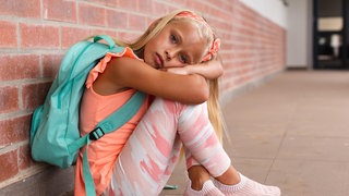 Grundschülerin sitzt traurig mit einem Rucksack in einem Schulkorridor.