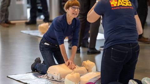 Eine Frau macht Herzdruckmassage an einer Puppe und schaut zu einer Rettungssanitäterin, die ihr Anweisungen zu geben scheint.