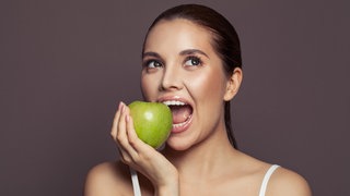 Junge Frau beißt in einen grünen Apfel. Anti Aging: Welche Lebensmittel sind gut gegen Hautalterung?