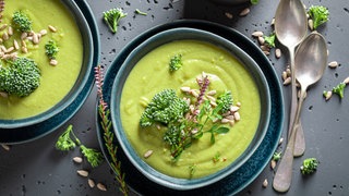 Grüne Brokkoli-Suppe in blauem Schälchen auf grauer Beton-Platte. Wird Brokkoli roh genossen, ist er besonders gesund. 