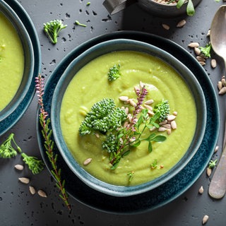 Grüne Brokkoli-Suppe in blauem Schälchen auf grauer Beton-Platte. Wird Brokkoli roh genossen, ist er besonders gesund. 