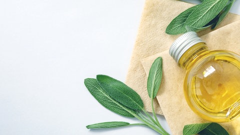 Eine kleine Glasflasche enthält Öl aus Salbei. Um die Flasche herum sind Salbeiblätter verteilt.