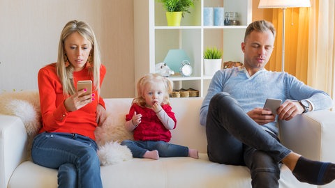 Kleinkind, Mann und Frau sitzen mit Handy in der Hand auf der Couch