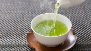 Grüner, dampfender Tee wird aus weißer Porzellankanne in weiße Tasse gegossen. Wie gesund sind grüner Tee und Matcha tatsächlich?