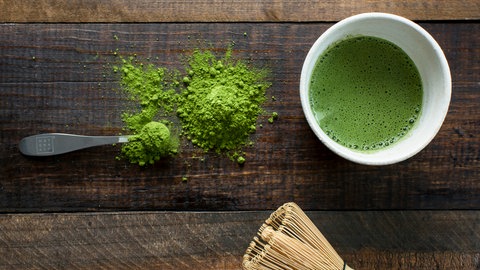 Leuchtend grünes Matchapulver auf Holzbrett, daneben zubereiteter Matcha in Schale und Besen. Wie gesund sind grüner Tee und Matcha tatsächlich?