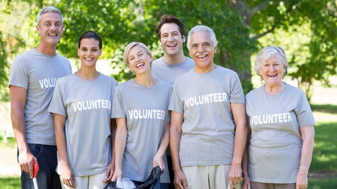 Lachende Menschen verschiedenen Alters tragen Shirts mit der Aufschrift "Volunteer" und halten sich im Arm. Wie sind Ehrenamtliche bei Unfällen oder Schäden versichert?