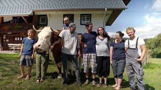 Ein Bauernhof im Schwarzwald, davor stehen 5 Erwachsene, 2 Kinder und eine Kuh