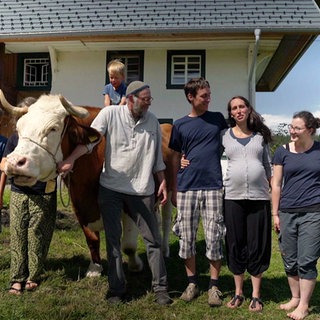 Ein Bauernhof im Schwarzwald, davor stehen 5 Erwachsene, 2 Kinder und eine Kuh