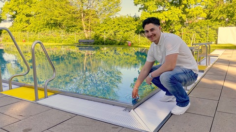 Der 17-jährige Mohamed kniet am Rand des Schwimmbeckens im Stettener Freibad und hält seine Hand ins Wasser. SWR1 Neuanfang. Mohamed: Flucht in ein neues Leben – ein junger Syrer schwimmt sich frei. Mohamed hat den syrischen Bürgerkrieg überlebt und fand eine neue Heimat in Stetten im Remstal. Warum ein Schwimmbad dabei entscheidend geholfen hat, erzählt er in SWR1 Neuanfang.