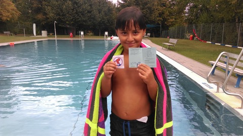 Mohamed im Jahr 2016, ein kleiner Junge steht am Rand eines Schwimmbeckens und zeigt seine Schwimmabzeichen. SWR1 Neuanfang. Mohamed: Flucht in ein neues Leben – ein junger Syrer schwimmt sich frei. Mohamed hat den syrischen Bürgerkrieg überlebt und fand eine neue Heimat in Stetten im Remstal. Warum ein Schwimmbad dabei entscheidend geholfen hat, erzählt er in SWR1 Neuanfang.