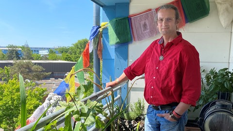 Neuanfang für Alexander nach einem Suizidversuch – Alexander steht auf einem mit buddhistischen Fähnchen geschmückten Balkon. Nach einer schweren, von Medikamenten ausgelösten Depression hat Alexander hat den Versuch einer Selbsttötung überlebt. Er sagt: "Suizidale Menschen möchten nicht sterben, sondern eigentlich nur anders leben". Deshalb hilft er Menschen in tiefen Lebenskrisen, deren Gedanken um Suizid kreisen.