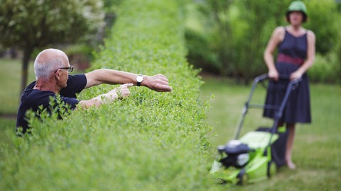 Ein Mann zeigt an einer Hecke auf seine Uhr waehrend seine Nachbarin den Rasen maeht.