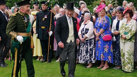 König Charles hat einen schwarzen Frack und einen Zylinder an und steht vor einer ausgewählte Menschenmegen. Begleitet wird er von einem älteren Herren in Uniform. 
