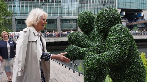 Königin Camilla streckt die Hand zwei Moos-Menschen entgegen. Sie lacht dabei und trägt einen hellen Trenchcoat.