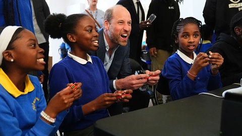 Prinz William sitzt mit einer Konosle in der Hand zwischen Schülerinnen und zockt freudig mit ihnen.