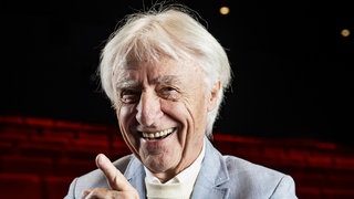 Emil Steinberger steht im leeren Theater in einem blauen Anzug und lacht in die Kamera