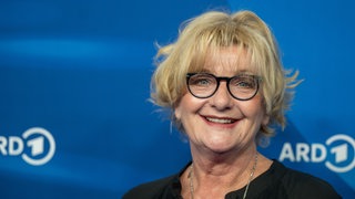 Die Schauspielerin Saskia Vester steht vor einem blauen Hintergrund und lacht in die Kamera.