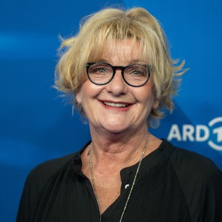 Die Schauspielerin Saskia Vester steht vor einem blauen Hintergrund und lacht in die Kamera.