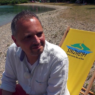 Philipp Dahlen sitzt auf einem Liegestuhl mit Aufdruck Sinzheim an einem See