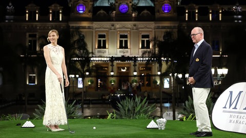 Links steht Fürstin Charlene in einem weißen langen Kleid und High Heels mit Golfschläger in der Hand, rechts steht Fürst Albert im schwarzen Sakko mit heller Hose und lächelt.
