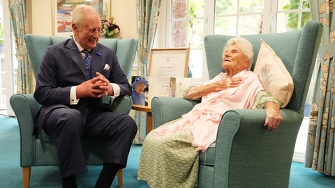 König Charles III. trifft die vermutlich älteste Person der Welt, die 116-jährige Ethel Caterham, im Hallmark Lakeview Care Home. Sie sitzen sich gegenüber. Charles lacht, Ethel schaut nach oben.