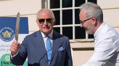 König Charles: Der Monarch hält ein großes Kuchenmesser in der Hand, trägt einen schicken Anzug und eine lässige Sonnenbrille dazu. Vor ihm auf dem Tisch steht eine Torte.