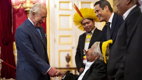 Ein weißhaariger älterer Mann im dunkelblauen Nadelstreifenanzug, König Charles, begrüßt lachend einen weißhaarigen Mann, der vor ihm im Rollstuhl sitzt, Chief Raoni Metuktire.