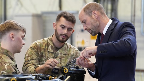 Ein Mann mit Halbglatze und im Anzug, Prinz William, hilft bei der Arbeit am Rotorkopf eines Helikopters.
