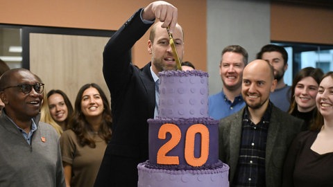 Vor einem jungen Mann, Prinz William, mit Halbglatze steht eine dreistöckige lila Torte mit einer orangefarbenen 20 drauf, die er mit einem großen Messer anschneidet.