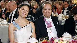 Prinzessin Victoria lächelt in weißem Kleid mit Krone neben ihrem Tischnachbarn Professor Susumu Ki-ta-gawa, einem der Gewinner des Nobelpreises für Chemie.