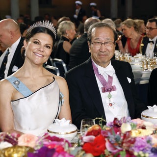Prinzessin Victoria lächelt in weißem Kleid mit Krone neben ihrem Tischnachbarn Professor Susumu Ki-ta-gawa, einem der Gewinner des Nobelpreises für Chemie.