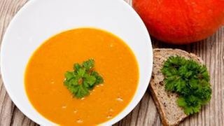 weiße Schüssel mit Kürbissuppe mit Petersilie als Deko, daneben eine Scheibe Brot und ein Kürbis