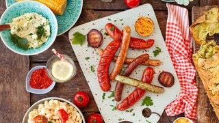 Die perfekten Beilagen zum Grillen: Draufsicht auf Kartoffelsalat, Nudelsalat, Zupfbrot und andere Beilagen, die um ein Schneidbrett mit Grillwürstchen platziert sind