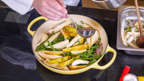 Über Birnen, Bohnen und Speck in der Pfanne wird etwas Brühe geschüttet. Das gibt dem Eintopf Würze.