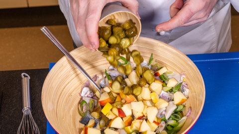 Die kleingeschnittenen Gewürzgurken werden dem Heringssalat zugegeben. Zum Schluss wird der Salat laut Rezept mit Pellkartoffeln angerichtet.