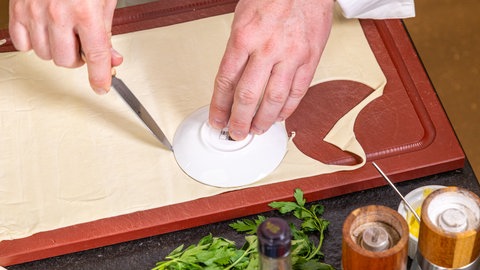 Mit einem kleinen weißen Schälchen schneidet der Koch auf einem braunen Brett für den Pilz-Pie runde Teigstücke aus dem Blätterteig und benutzt dabei ein Messer. Im Vordergrund stehen Pfeffer- und Salzstreuer und ein Bund Petersilie liegt dahinter.