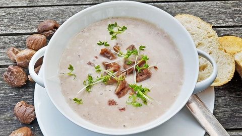 Cremige Maronensuppe: Eine Maronensuppe mit Stückchen von Maronen und Kräutern darüber gestreut ist in einer weißen Suppenschale angerichtet. Auf dem Unterteller liegen ein Suppenlöffel und Scheiben von Weißbrot. Links daneben liegen mehrere Maronen.