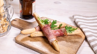 Die selbstgemachten Bierstängel liegen mit Schinken und Ruccola umwickelt auf einem Holzbrett. Daneben steht ein Bierkrug mit weiteren Bierstangen und eine flasche Bier.