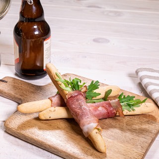 Die selbstgemachten Bierstängel liegen mit Schinken und Ruccola umwickelt auf einem Holzbrett. Daneben steht ein Bierkrug mit weiteren Bierstangen und eine flasche Bier.