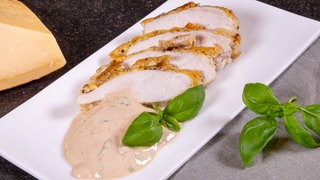 Die Geflügelbrust in Panade aus Parmesan ist in Scheiben geschnitten und zusammen mit dem Dip auf einem weißen Teller angerichtet. 