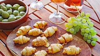 Holzbrett mit Käsehörnchen, daneben Trauben und ein Glas Rose-Wein