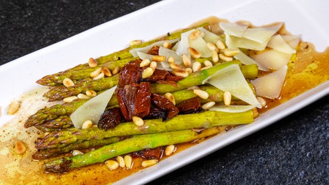 Eine weiße Platte mit grünem Spargel, auf dem Pinienkerne verstreut sind, zwischen den Spargelstangen liegen getrocknete Tomaten und oben drauf gehobelter, geschmolzener Parmesan